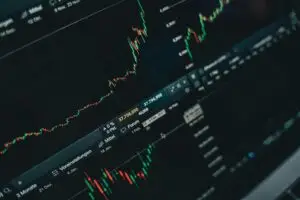 Close-up view of a computer screen displaying live stock market charts with green and red candlestick patterns and price movements, showing financial trading data and technical analysis indicators on a dark background.